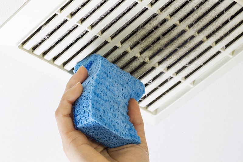 Close up horizontal photo of female hand cleaning dirty bathroom fan vent cover with blue sponge.