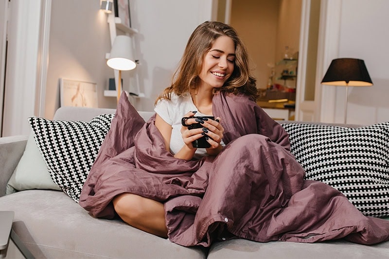 A Guide to Indoor Humidity Control. Photo of a young woman enjoying a hot beverage on her couch wrapped in a blanket.